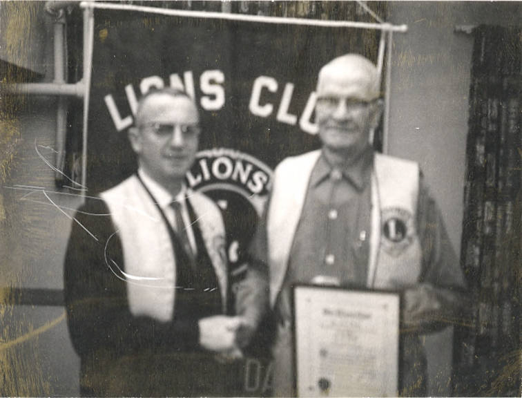 Two men are standing in front of a sign that reads "Lions Club." They are wearing vests with a logo, and one man is holding a certificate. They appear to be shaking hands.