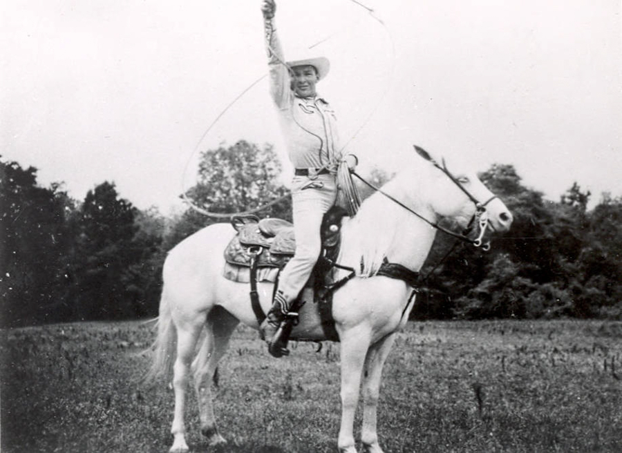 A person in cowboy attire is riding a white horse in an open field. The person is holding a lasso in the air with one hand and is smiling. The background shows trees and grass.