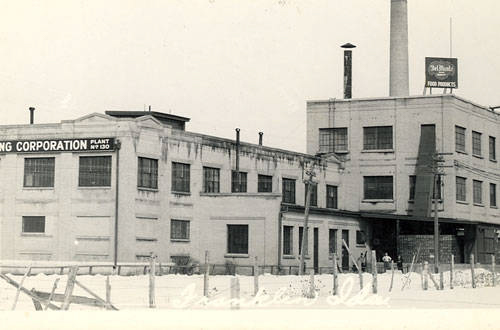 An industrial building with multiple sections and large windows, featuring signage on the left side that reads "Corporation Plant No. 320." A tall chimney is visible, and on the top right corner of the building, a sign reads "Bell Brand" and "Food Products." A fence runs along the front of the building.