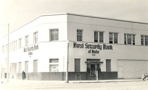 A two-story building on a street corner with signage that reads "First Security Bank of Idaho N.A." A person is walking on the sidewalk in front of the building. Several windows display "First Security Bank of Idaho N.A." Signs are visible on both sides of the building. A bicycle is parked near the entrance.