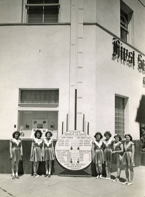 Seven women in matching outfits stand in front of a building. They are positioned beside a vertical sign with numbers and labels related to different military branches: Army, Navy, Air Corps, and Marines, with respective monetary goals. At the bottom, it reads "FRANKLIN CO. QUOTA" and asks, "HAVE YOU BOUGHT YOUR BOND TODAY?" The sign tracks financial contributions toward a quota of $275,000. The building has a window with some objects displayed and a partial sign with the word "Music" visible.