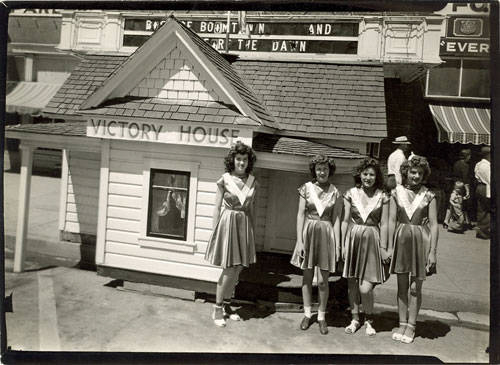 Four young women are standing in front of a small house with a sign reading "Victory House." They are wearing matching uniforms with skirts and sandals. In the background, there is a building with awnings and partially visible text that includes "BOOM TOWN AND" and "THE DAWN." Passersby can be seen walking nearby.