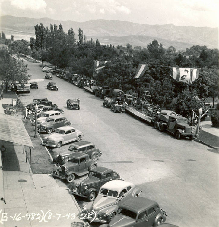 Parked cars line a street, with several vehicles decorated as parade floats on the opposite side. Trees and mountains are visible in the background. A few flags hang above the floats. Handwritten text reads "G-16-482 (8-7-43-2P)" at the bottom.
