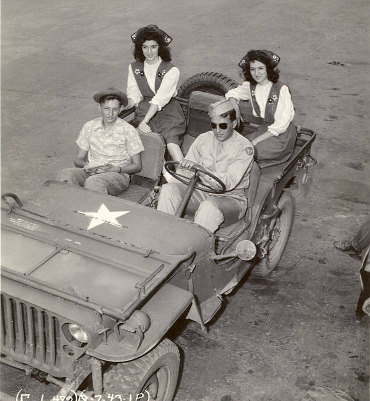 A group of four people in a military jeep. Two women are seated in the back, both wearing matching outfits with vests and bows in their hair. In the front, a man in a military uniform with sunglasses is driving, and next to him, a man in a patterned shirt sits in the passenger seat. The jeep has a visible star on the hood.