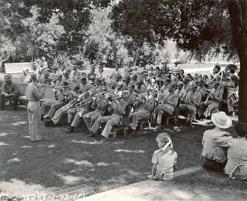 A band composed of many musicians is seated on benches in a shaded outdoor area, playing various brass instruments. A conductor stands in front of them, leading the ensemble. To the side, a child sits on the grass with her back to the band, watching an adult couple seated nearby. Dense trees and bushes form a natural backdrop. The bottom left corner has handwritten numbers and letters.