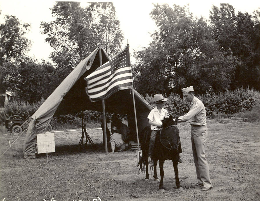 A person is on horseback next to a man in a uniform. They are outside near a large tent displaying an American flag. There are people sitting under the tent. Trees and a vehicle are in the background.