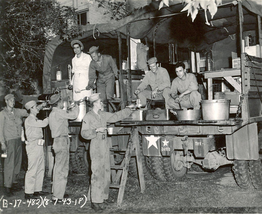A group of men in military uniforms are gathered around a mobile field kitchen. Two men stand inside the truck serving food from large pots, one with a ladle. Another man, dressed in a white uniform and hat, supervises the serving. Several men stand on the ground, holding cups and plates to receive food. The vehicle has a star symbol and the number 24571 visible on its side. There is a ladder for access to the truck's serving area. In the lower left corner, the text "G - 17 - 482) (8 - 7 - 43. 1P)" is visible.