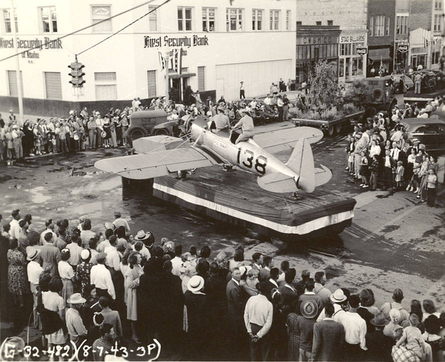 A crowd of people is gathered around a platform in the street, where a small aircraft with the number 138 is displayed. A person is seated in the cockpit of the aircraft. The adjacent building is marked First Security Bank. Numerous onlookers surround the scene, some dressed in hats. Urban buildings and a traffic light can be seen in the background, along with parked cars. Text on the image includes "G-32-482," "8-7-43-3P."