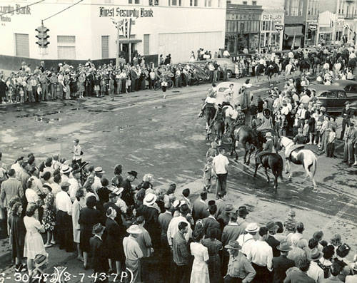 A large crowd stands along a street watching a parade. Several people on horseback ride down the street. The audience includes men, women, and children, many wearing hats. Across the street is a building labeled "First Security Bank," with more spectators gathered in front. Additional shops and buildings are visible in the background.