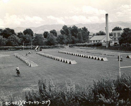 Open field with rows of small tents arranged neatly. A person on horseback is visible in the foreground. A building with a tall chimney stands in the background, surrounded by trees. Text at the bottom reads "(8-7-43-3P)".