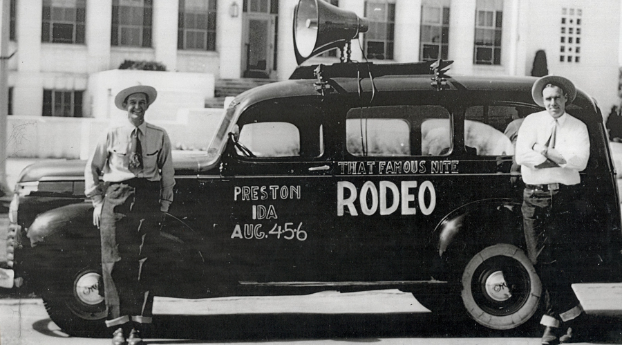 Two men wearing hats are standing next to a vehicle with a large speaker mounted on top. The vehicle has text on the side that reads "THAT FAMOUS NITE RODEO" and "PRESTON IDA AUG. 4 5 6". A building is visible in the background.