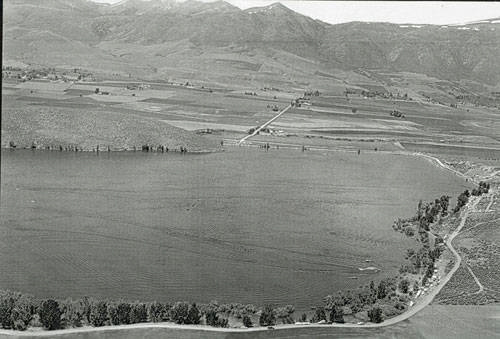 An aerial view of a large body of water surrounded by rural land. A road runs alongside the water, bordered by trees. Farmland stretches out towards distant hills and mountains in the background.