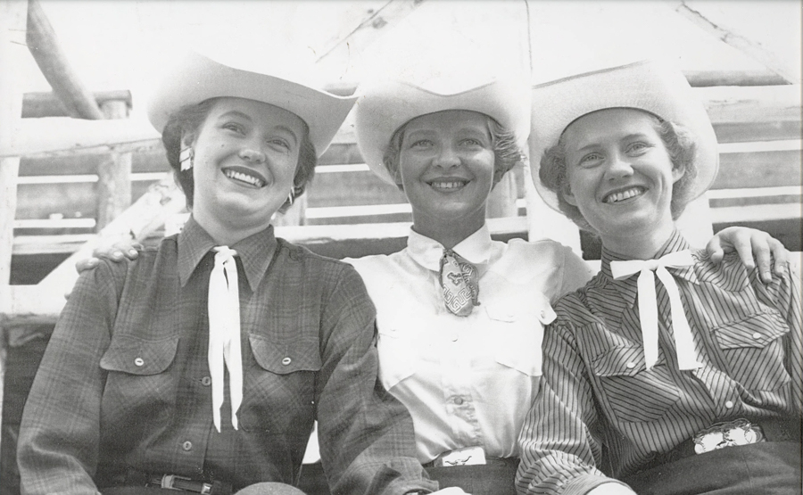 Three women are sitting side by side, each wearing a cowboy hat and western-style shirts. They are smiling, with their arms around each other. The background shows part of a wooden structure.