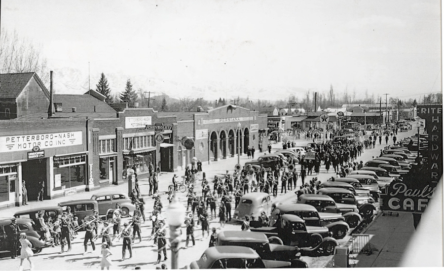 A bustling street scene with a parade featuring numerous people walking, some playing musical instruments. The street is lined with buildings, including the Petterborg-Nash Motor Co. Inc. and a Chevrolet dealership. Numerous cars are parked along the road. Signs for Paul's Cafe and Rite Hardware are visible. People watch the parade from the sidewalks. The background shows trees and distant hills.