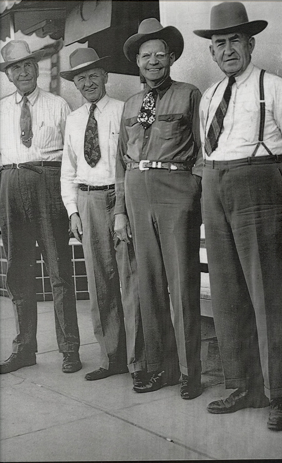 Four men standing side by side on a sidewalk. They are wearing hats, dress shirts, and ties. Each man is smiling, and their hands are relaxed at their sides. There is a building wall and a ledge visible in the background.