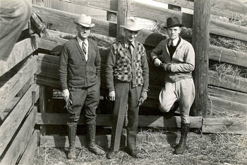 Three men are standing in front of a wooden structure, possibly a fence or corral. Two men wear hats and cardigans, while the third wears a sweater and leans against the fence. They are dressed in early 20th-century clothing, with each man wearing boots.