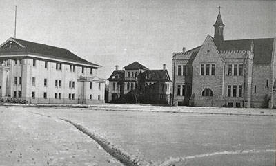 Three buildings are positioned around an open area. The building on the left has a flat roof and multiple windows. The center structure is smaller, featuring a more intricate design. The building on the right is large, with a peaked roof and a small tower. The ground appears to be covered in snow or frost.