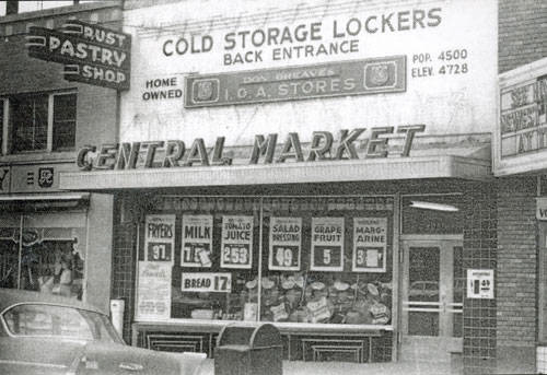 The image shows the exterior of a building labeled "Central Market" with a sign advertising "Cold Storage Lockers Back Entrance." Above the main signage, it mentions "Home Owned I.G.A. Stores." There are several signs displaying prices for products such as fryers, milk, tomato juice, salad dressing, grapefruit, margarine, and bread. To the left, there is a sign for a pastry shop. A vintage car is parked in front of the market. Nearby, there's a small sign with numbers "4500" and "4728," possibly indicating population and elevation.