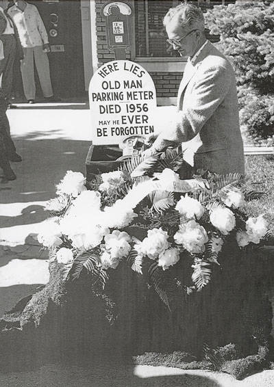 A man stands beside a burial site covered in flowers. A sign reads, "Here lies Old Man Parking Meter, Died 1956, May he ever be forgotten." There are a few people and a building visible in the background.