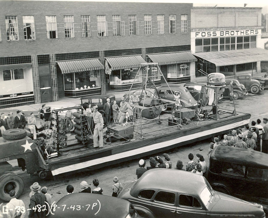 A street scene featuring a parade float displaying various machinery being viewed by a crowd. Several people are standing on the float, interacting with the equipment. The surrounding area has cars parked on the street and people watching the scene. A building in the background displays a sign reading "FOSS BROTHERS." Additional text includes a date and reference number: (8-7-43-7P) and [133-482].