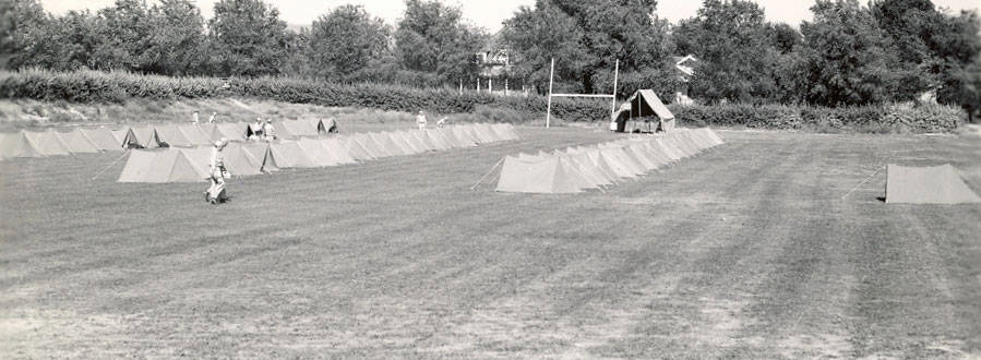 A field with numerous small tents arranged in rows. Several people are walking or standing among the tents. There are trees and hedges in the background, and a larger tent near goalposts is visible.