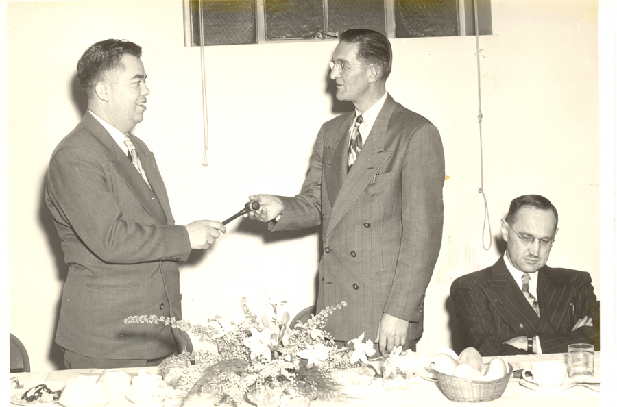 Two men in suits are standing and exchanging a gavel. Another man in a suit is sitting at a table, looking down, with his arms crossed. The table has a floral arrangement, a basket of rolls, a cup, and a glass. A window and a pull cord are visible in the background.
