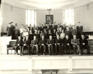A group of people arranged in three rows inside a large room with high windows. The first row is seated, while the two rows behind are standing. They are dressed in formal attire, and there is a framed portrait on the back wall above them. The room appears to be a hall or auditorium with wooden paneling.
