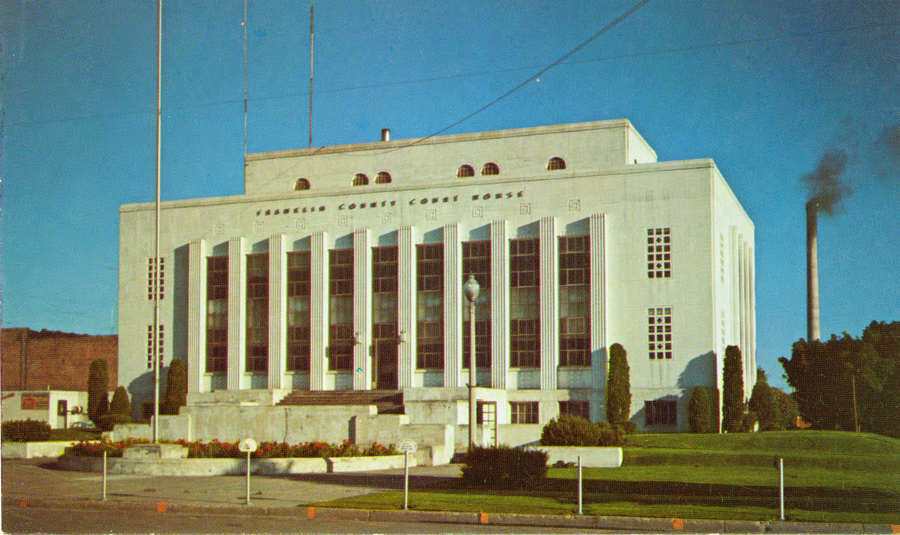 A large building with tall columns and a series of large windows stands prominently. The words "Douglas County Court House" are visible on the front. There is a manicured lawn and several tall trees around the structure. A flagpole and a street lamp are in front. In the background, a smokestack with smoke coming out is visible.