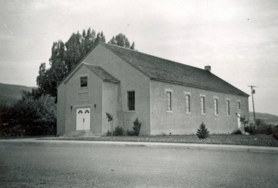 A rectangular building with a gabled roof sits next to a road. The facade features two doors with an arched design above them, and several tall windows line the side of the building. There are trees and small bushes around the building, and a utility pole is visible in the background.