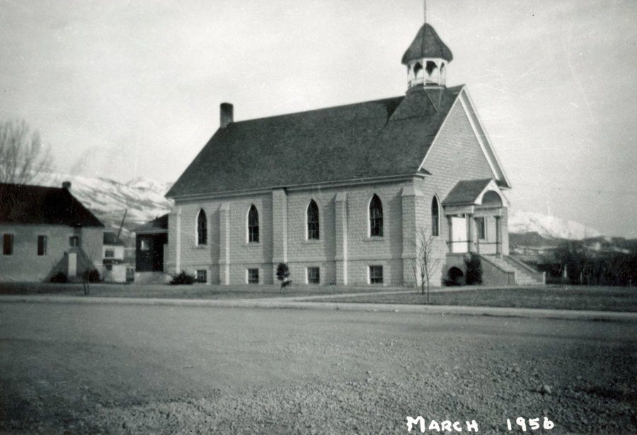 A church with arched windows and a steep roof stands next to a smaller building. The church has a prominent entrance with stairs leading up to it and a bell tower on top. Mountains can be seen in the background. The words "March 1956" are written in the lower right corner.