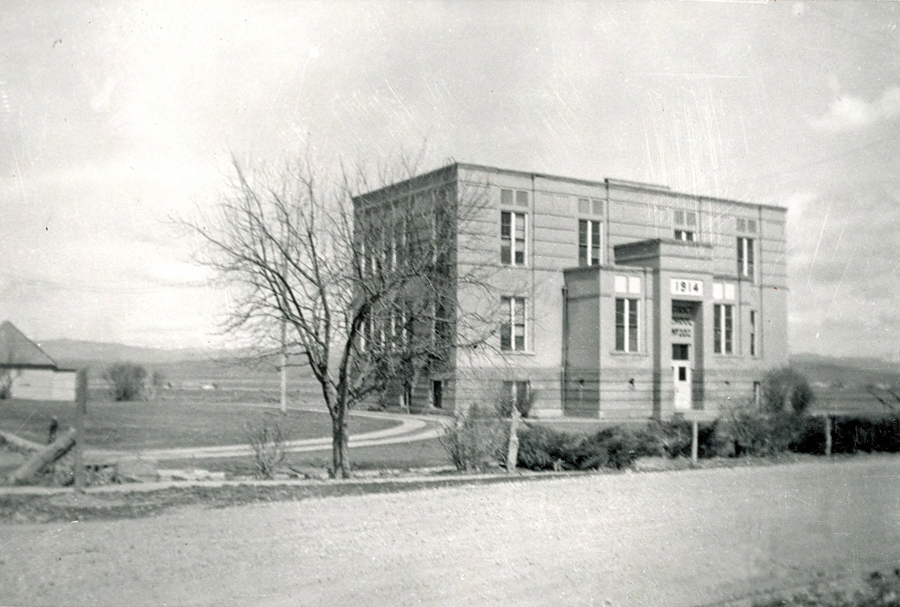 A large rectangular building stands in a rural setting, featuring multiple windows and a protruding entrance. The entrance has the numbers "1914" displayed above it, along with the word "SCHOOL" above the doorway. A leafless tree is situated in the foreground, and a dirt road runs parallel to a fence in front of the building. In the background, there are open fields and distant hills.