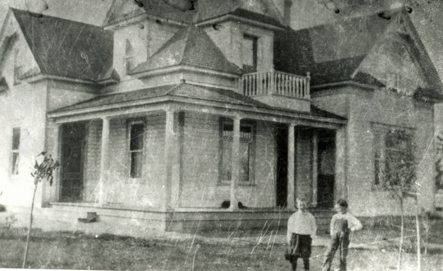 Two children stand in front of a large house with a porch and multiple roof peaks. The house has a balcony above the porch, and there are small trees in the yard.