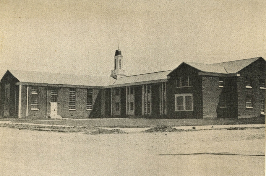 A large brick building with multiple windows and a small cupola on the roof. The structure features a central section with a row of tall columns and two symmetrical wings. The foreground shows a dirt area, possibly undeveloped land or a construction site.