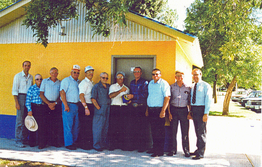 A group of thirteen men standing in a line outside a yellow and blue building. Some are holding hats, and one man is holding a small object in his hand. There are trees and parked cars in the background.