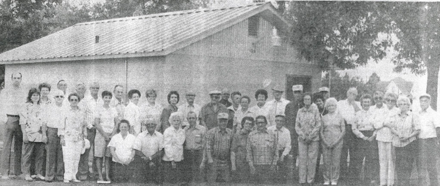 A group of people standing in front of a building with a sloped roof. There are trees visible in the background. The individuals are posed in a line, facing forward. No text is present.