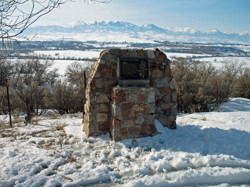 A stone monument with a plaque stands on a snowy landscape. In the background, there are trees and a view of distant mountains. The plaque contains text but is not legible from this angle.
