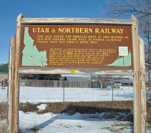 A large wooden sign in a snowy landscape. The sign reads: "UTAH & NORTHERN RAILWAY. One mile south and directly west of this highway an old 1878 railway grade, still is visible although trains have not used it since 1880." There are two map illustrations on the sign, with the numbers "91" and "257" displayed. The text continues with historical information about Jay Gould, Hamilton, McCammon, and the Oregon Shortline, noting specific years and their roles in railway expansion. A building and bare trees are visible in the background.