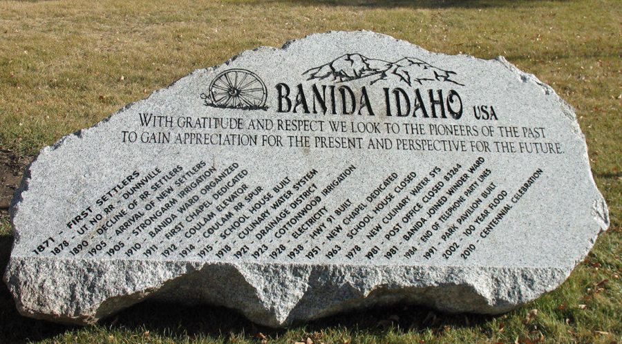 A large stone monument is engraved with the text "BANIDA IDAHO USA." An illustration shows mountains and a wagon wheel. The inscription reads, "With gratitude and respect we look to the pioneers of the past to gain appreciation for the present and perspective for the future." Below, a timeline lists key historical events from 1877 to 2010, including the arrival of settlers in 1895, the building of Banida Ward in 1913, and the centennial celebration in 2010.