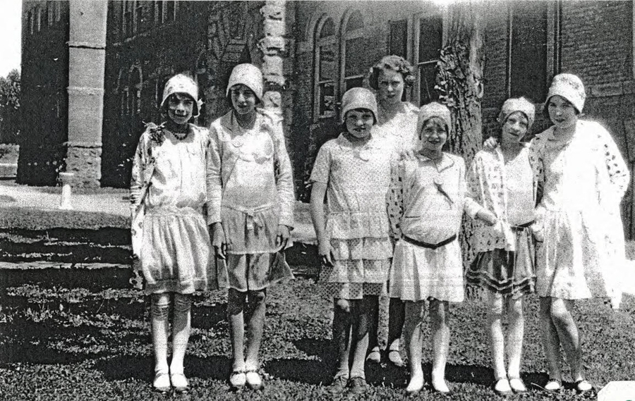 A group of eight young girls standing on grass in front of a large building. They are wearing dresses and cloche hats, typical of early 20th-century fashion. The building behind them has arched windows and stone pillars.