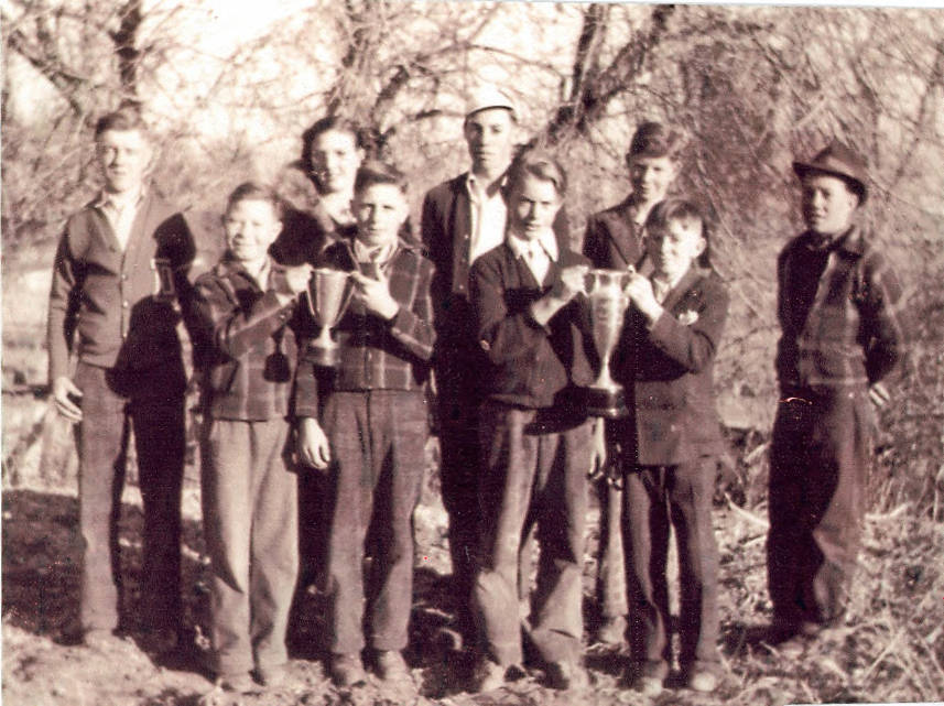 A group of nine people standing outside in a natural setting. Four young boys are holding two large trophies. Four adults and one young person stand behind and beside them, watching and smiling. The individuals are dressed in casual clothing, and there are leafless trees in the background.