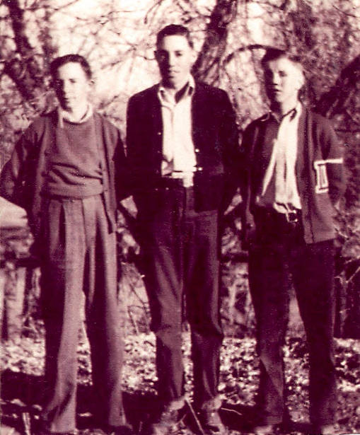 Three young men standing outdoors in front of trees. They are dressed in casual clothing, with the one on the right wearing a jacket with a letter patch on the sleeve. They stand with their arms by their sides and look towards the viewer. The background shows branches and foliage.