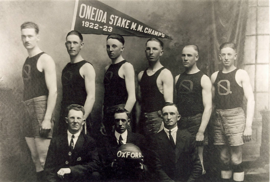 A group of eight men posed for a portrait, with five standing in the back and three seated in front. The standing men wear sleeveless shirts and shorts, while the seated men are in suits. Above, a pennant reads "ONEIDA STAKE M.M. CHAMPS 1922-23." The word "OXFORD" is on a ball held by one of the seated men.