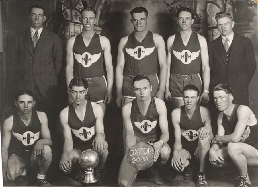 A group of nine men pose for a team photograph. Seven wear matching athletic uniforms with "M M" logo featuring wings. Two men dressed in suits stand on either side. One man in the front holds a ball labeled "Oxford 30-31." Another man in the front has a trophy in front of him. They are in an indoor setting with a decorative backdrop.