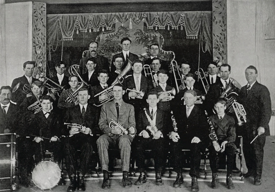 A group of men and boys, arranged in rows, holding musical instruments like trumpets and trombones. They are dressed in suits and ties, and some wear bow ties. The backdrop features ornate drapes and a decorative pattern. A drum is visible at the front of the group.