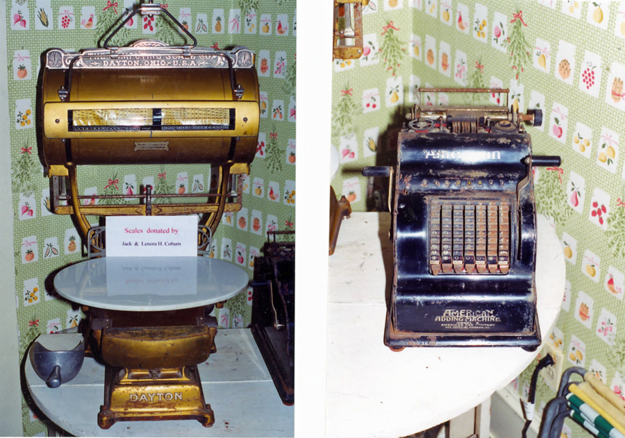 On the left, a brass weighing scale with ornate details is displayed. The scale is labeled "Dayton, Ohio, U.S.A." and has a round white platform. A card below reads "Scales donated by Jack & Lenora H. Coburn." On the right, a black adding machine with multiple keys and the label "American Adding Machine" rests on a round table. The background features patterned wallpaper with fruit and floral designs.