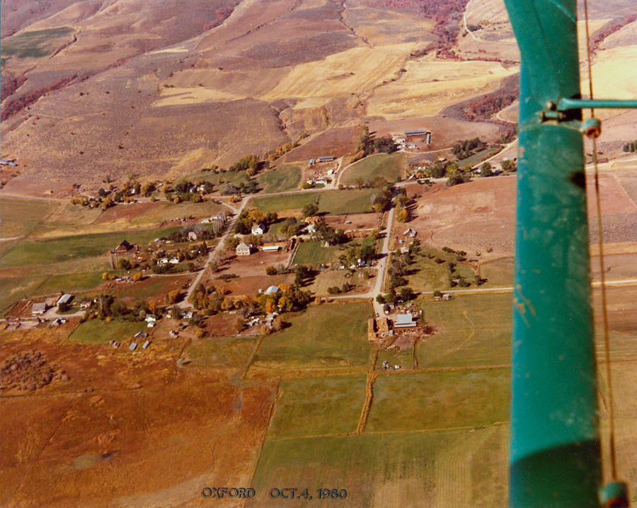 Aerial view of a rural landscape showing a small town with several buildings scattered along a grid layout of streets. There are open fields surrounding the town, with patches of farmland. A large green structure partially obscures the right side of the image. The text at the bottom reads "OXFORD OCT. 4, 1980."