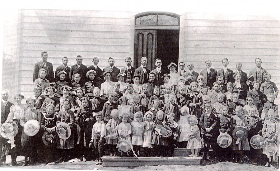 A large group of people, including men, women, and children, is gathered in front of a wooden building with a central door. The adults are mostly dressed in formal attire, while the children hold hats and wear light dresses or suits. The group is arranged in several rows, with the children in front and adults behind.