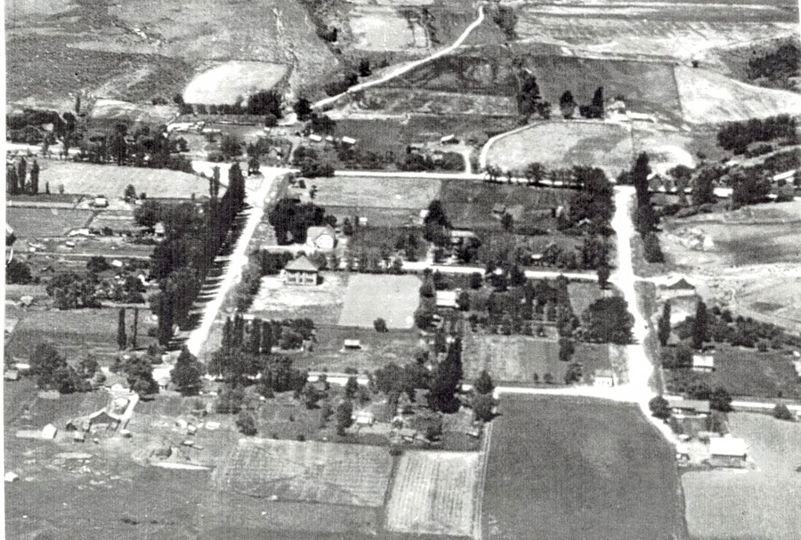 An aerial view of a rural landscape with a grid of roads and scattered houses. The area includes rectangular fields, clusters of trees, and various buildings. There is no visible text.
