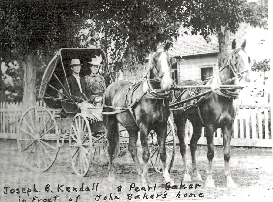 A man and a woman are seated in a horse-drawn carriage. The carriage is pulled by two horses and is positioned in front of a house. The man is wearing a suit and a hat, and the woman is in a dress with a hat. There are trees and a picket fence in the background. Text reads: "Joseph B. Kendall & Pearl Baker in front of John Baker's home."