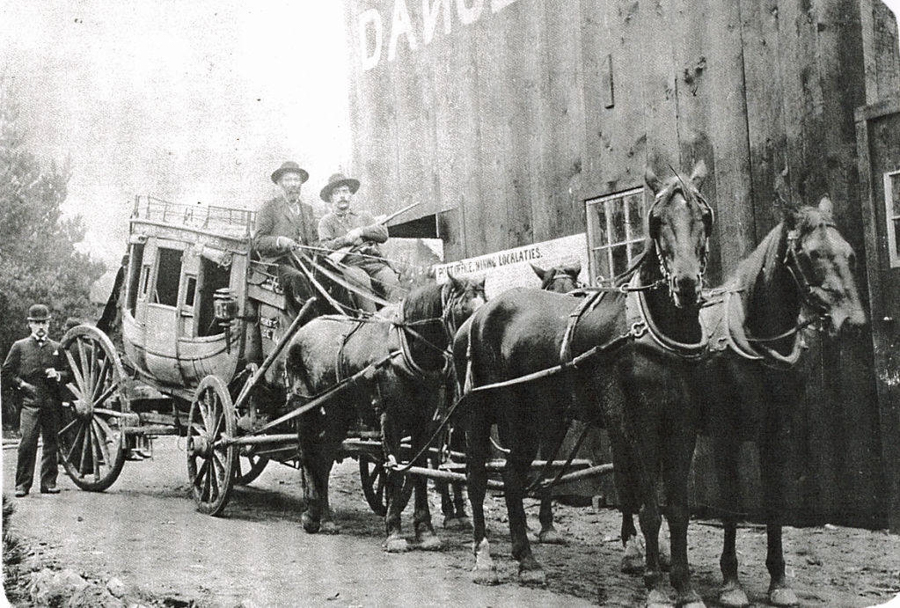 Two men are seated on a horse-drawn stagecoach, with three horses at the front. A man in a suit and bowler hat stands beside the horses. There is a large wooden building in the background with a sign partially visible. Another sign reads "THE MAIL OF THE MINING LOCALITIES."